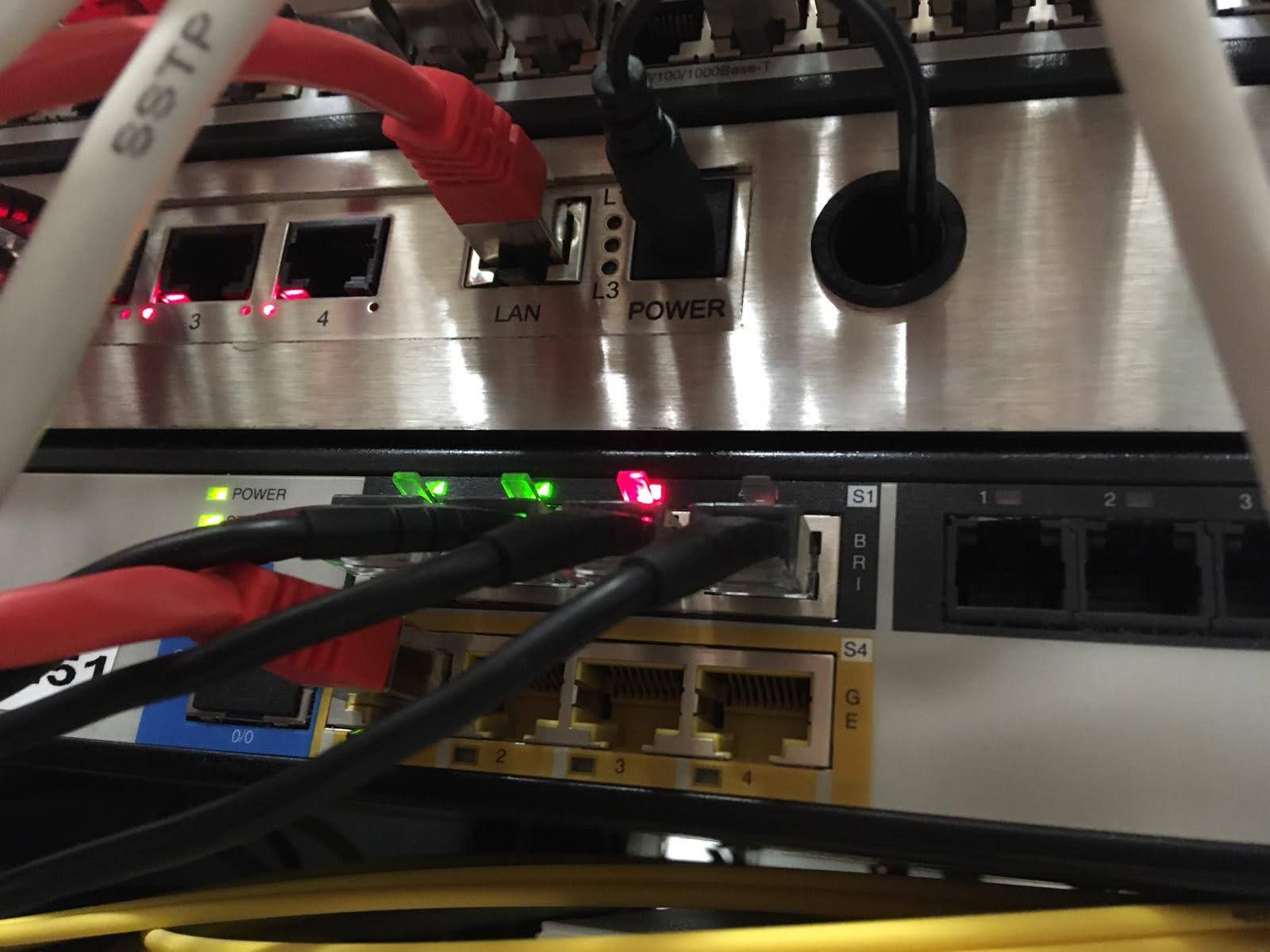 Rückseite von Netzwerkgeräten im Schrank, angeschlossene Patchkabel und ein Anschlussfeld mit ISDN-BRI-Ports.